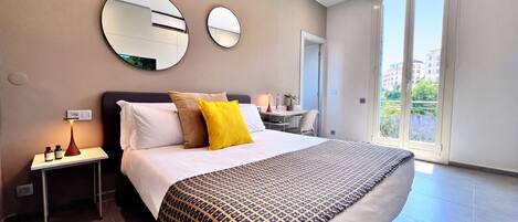 A modern hotel room with a large bed, two bedside tables, a desk, and a balcony with a city view.