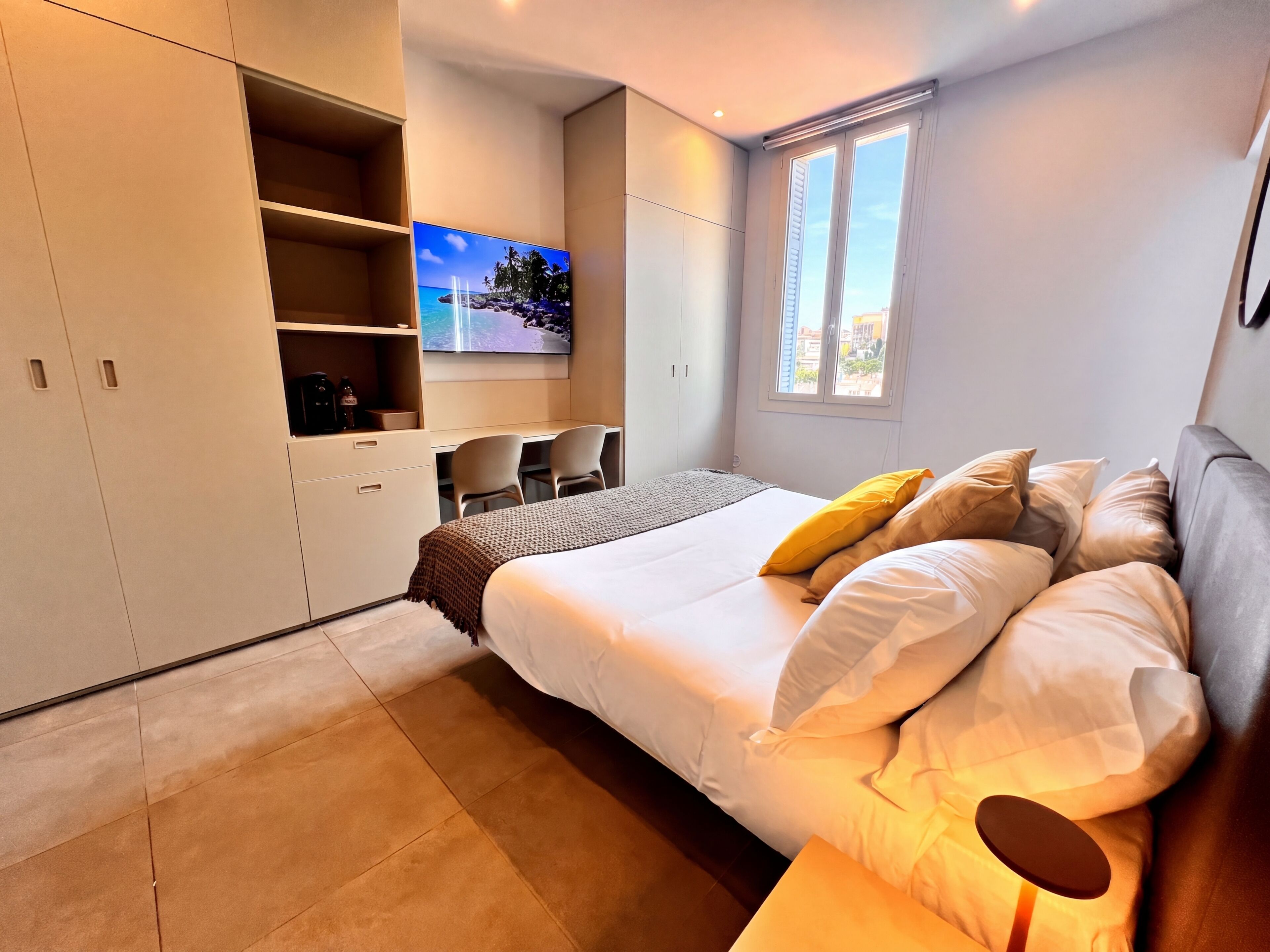 Een moderne slaapkamer met een groot bed, een tv aan de muur, een bureau met stoelen, en een raam met uitzicht.