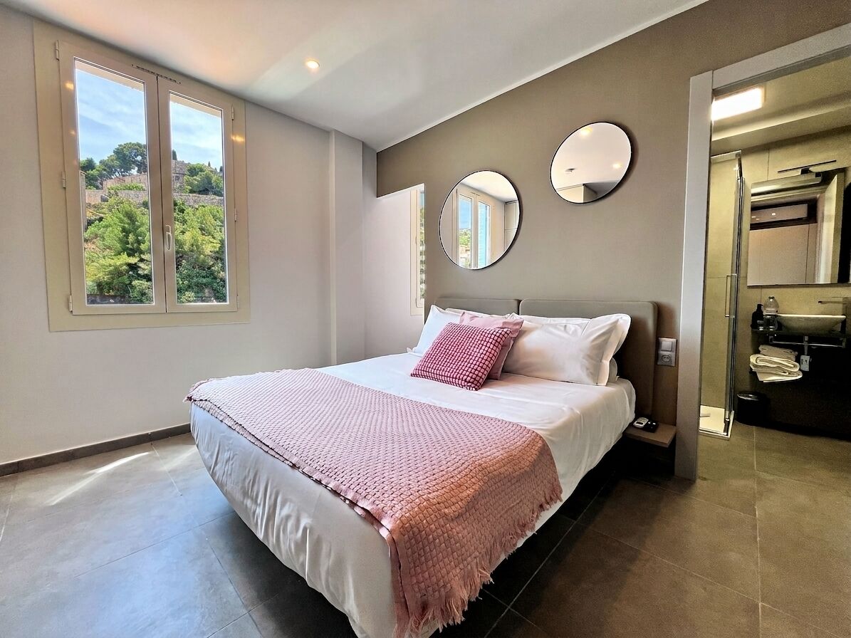 A bedroom with a bed, two bedside mirrors, a window, and a view of greenery.