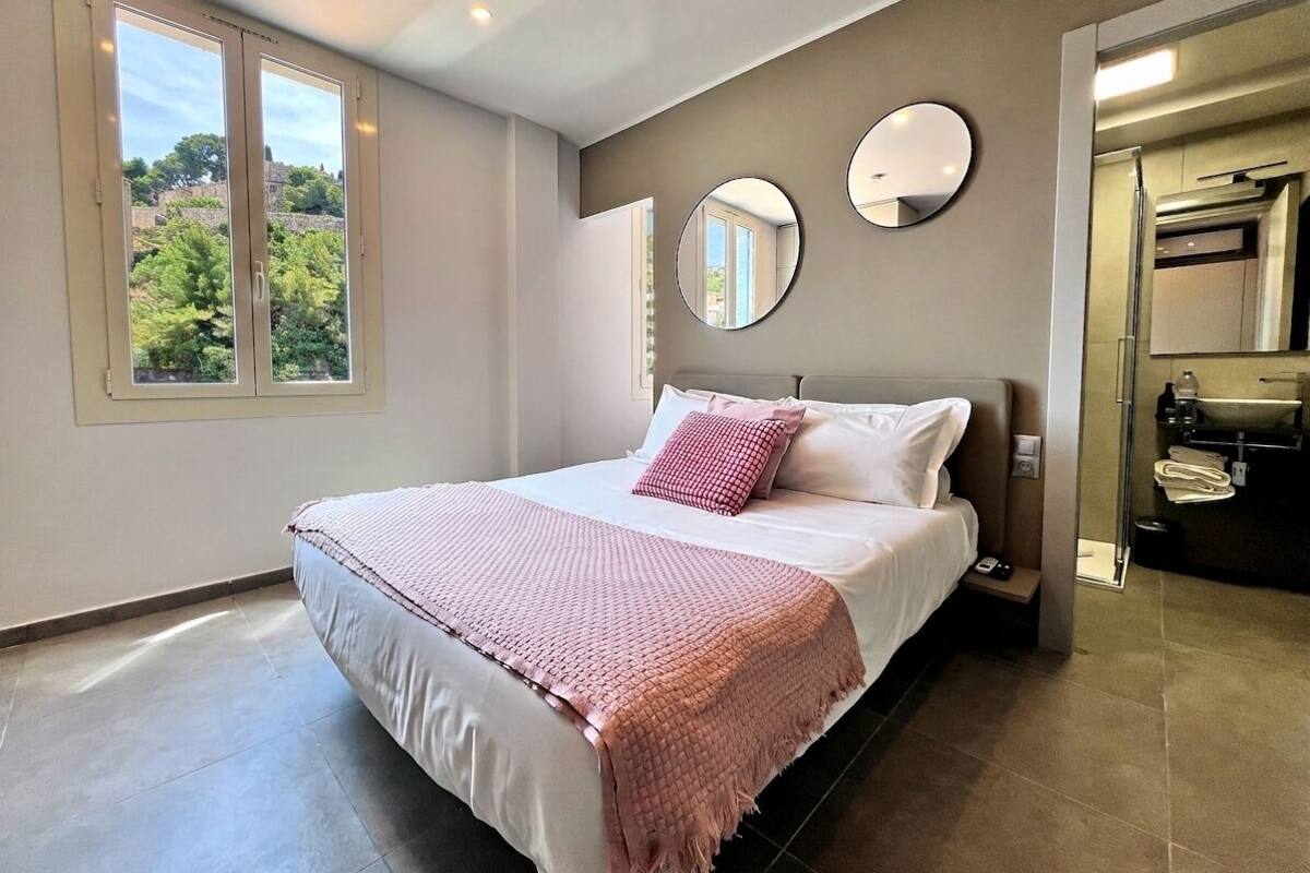 A bedroom with a bed, two bedside mirrors, a window, and a view of greenery.
