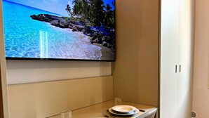 Un comedor con una televisión mostrando una escena de playa, una mesa dispuesta con platos y cubiertos, y una silla.