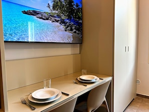 Un comedor con una televisión mostrando una escena de playa, una mesa dispuesta con platos y cubiertos, y una silla.