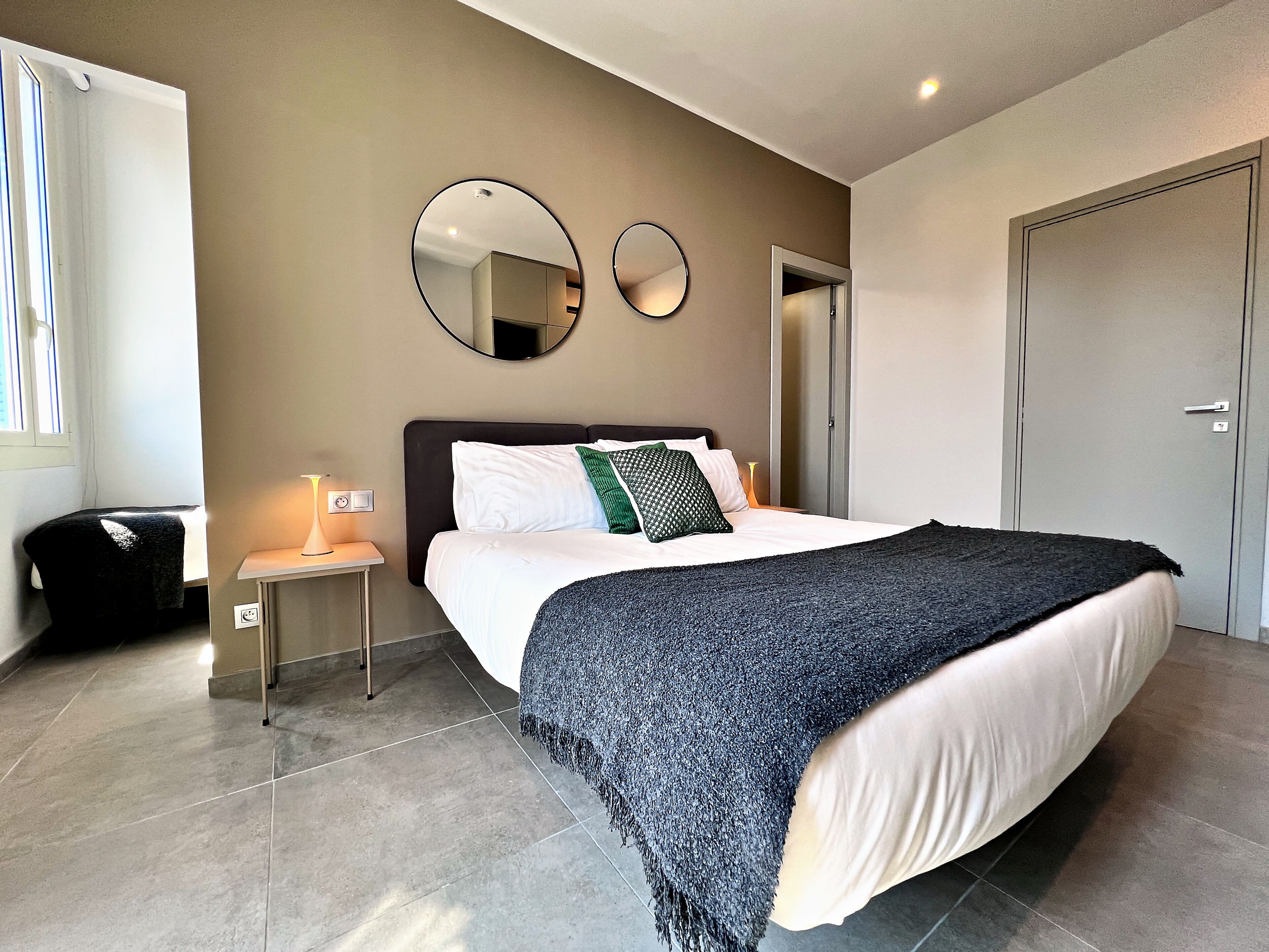 A bedroom with a bed, bedside table, lamp, two wall mirrors, and a wardrobe.