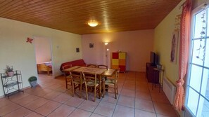 Ein Zimmer mit Holzboden, Fliesenboden, einem Esstisch mit Stühlen, einer Couch, einem Fernseher und einem Fenster mit Vorhängen.