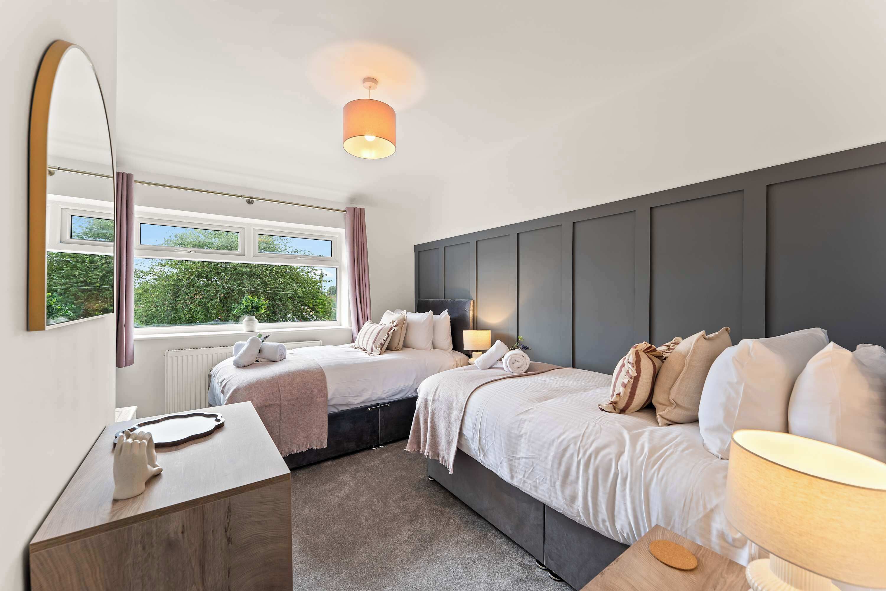 A modern bedroom with two beds, a large mirror, a desk, and a window with a view of greenery.