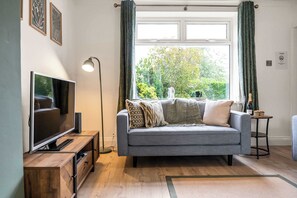Ein Wohnzimmer mit grauem Sofa, einem Flachbildfernseher auf einem Holzständer, einer Stehlampe und einem Fenster mit Vorhängen.