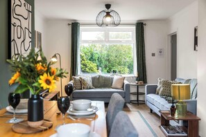 Ein Wohnzimmer mit einer Couch, einem Esstisch mit Tellern und Gläsern, einer Vase mit Blumen und einem Fenster mit Vorhängen.