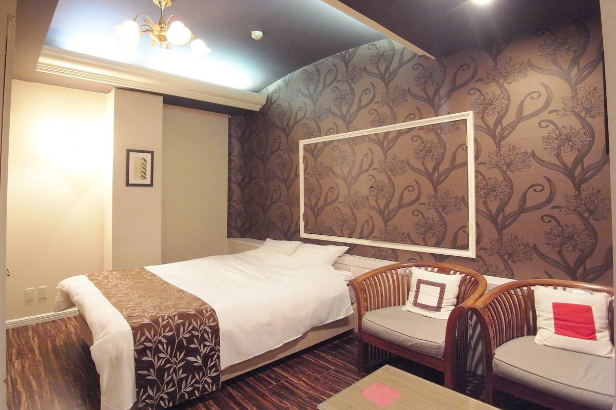 A hotel room with a bed, two chairs, a table, a ceiling light, and a wall with a floral pattern.