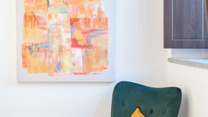 Un fauteuil de couleur bleu sarcelle agrémenté d’un coussin jaune moutarde, un tabouret en bois et un grand tableau abstrait accroché au mur.