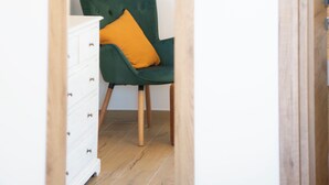 Une pièce avec un plancher en bois, un fauteuil vert recouvert de tissu et agrémenté d’un coussin orange, et une commode blanche.