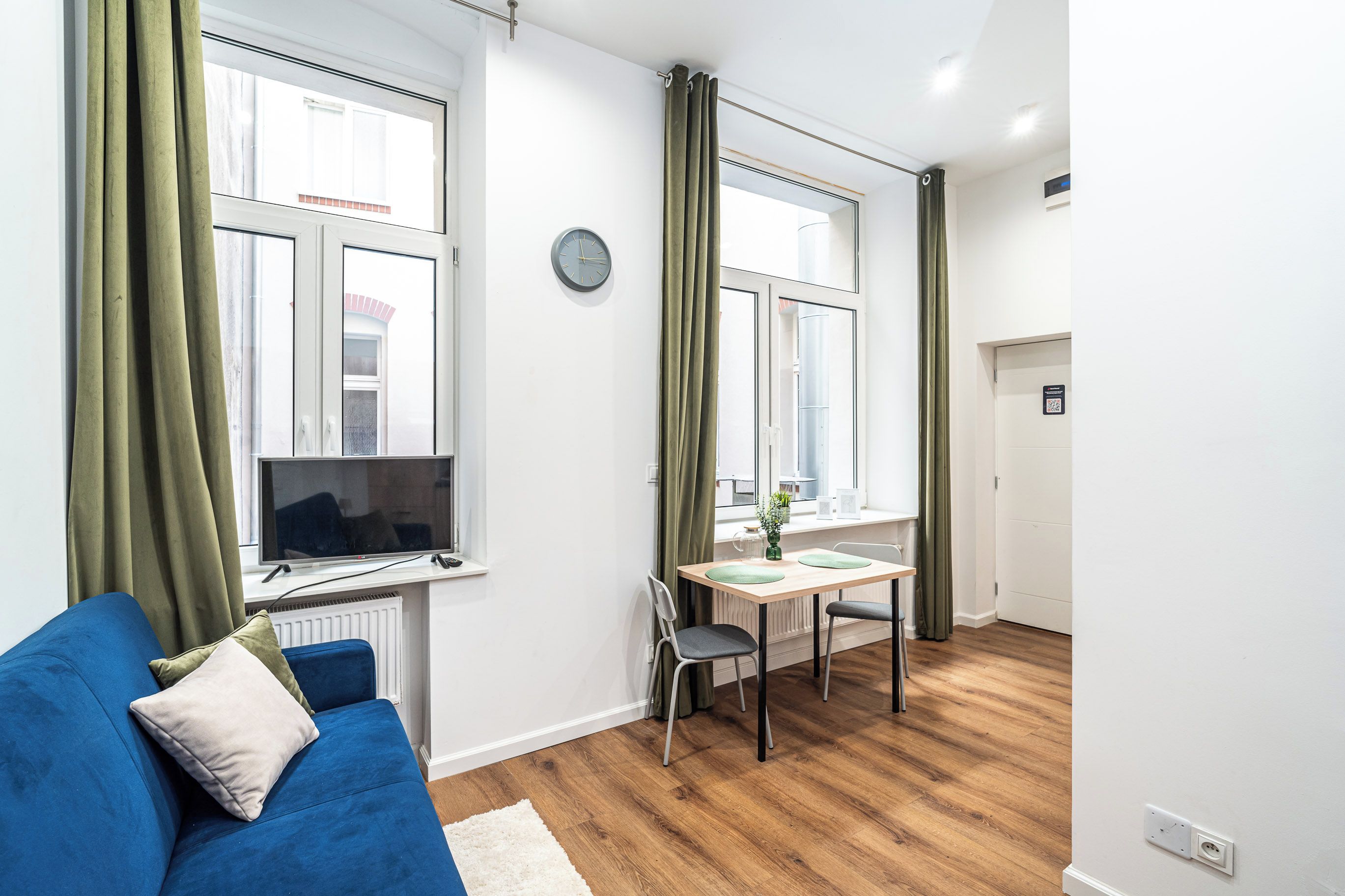 Ein modernes Apartment mit einem blauen Sofa, einem Esstisch mit Stühlen, einem Fernseher und einem großen Fenster mit Vorhängen.