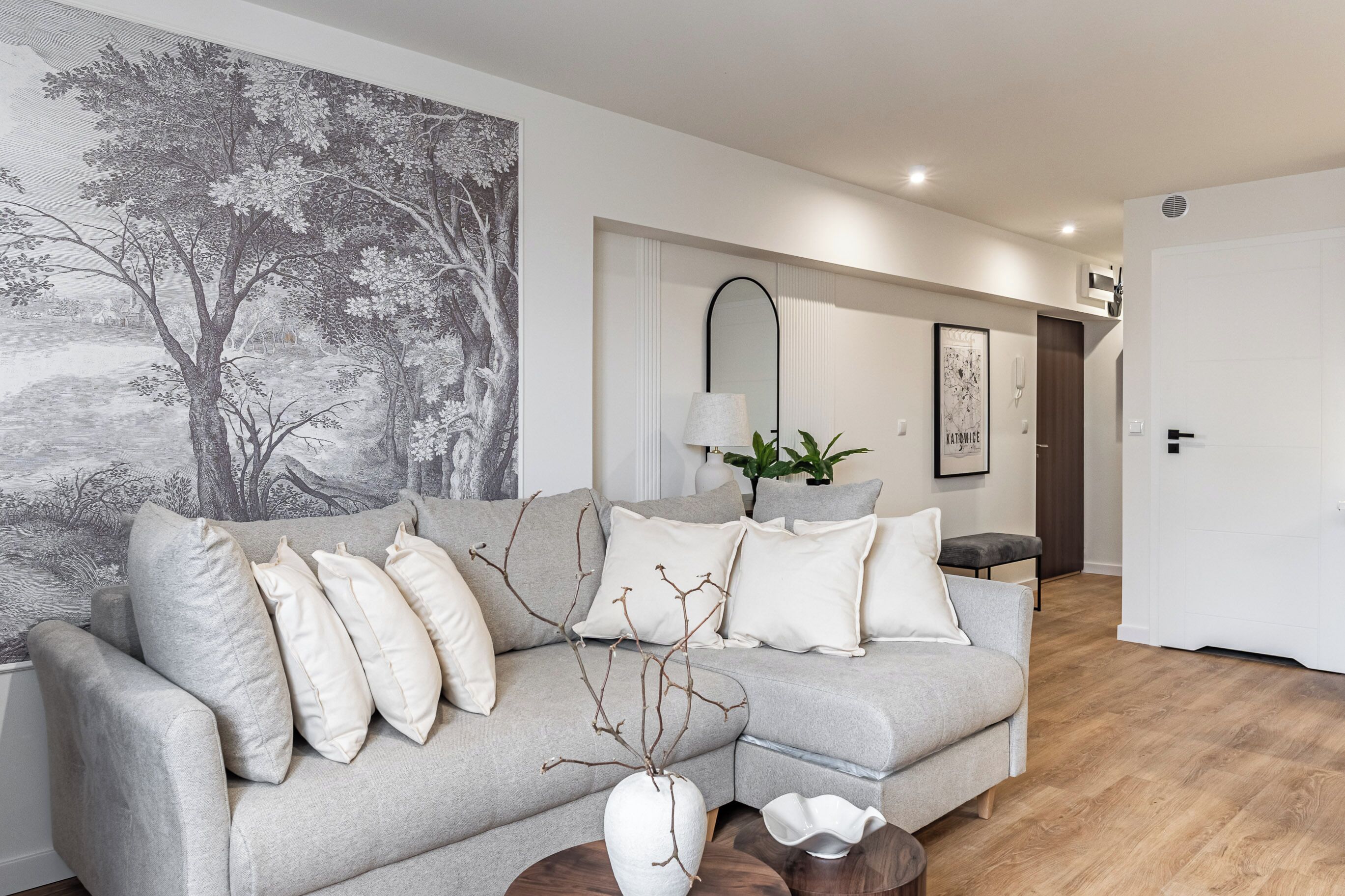 A modern living room with a grey sofa, a large wall mural, a mirror, and a plant.