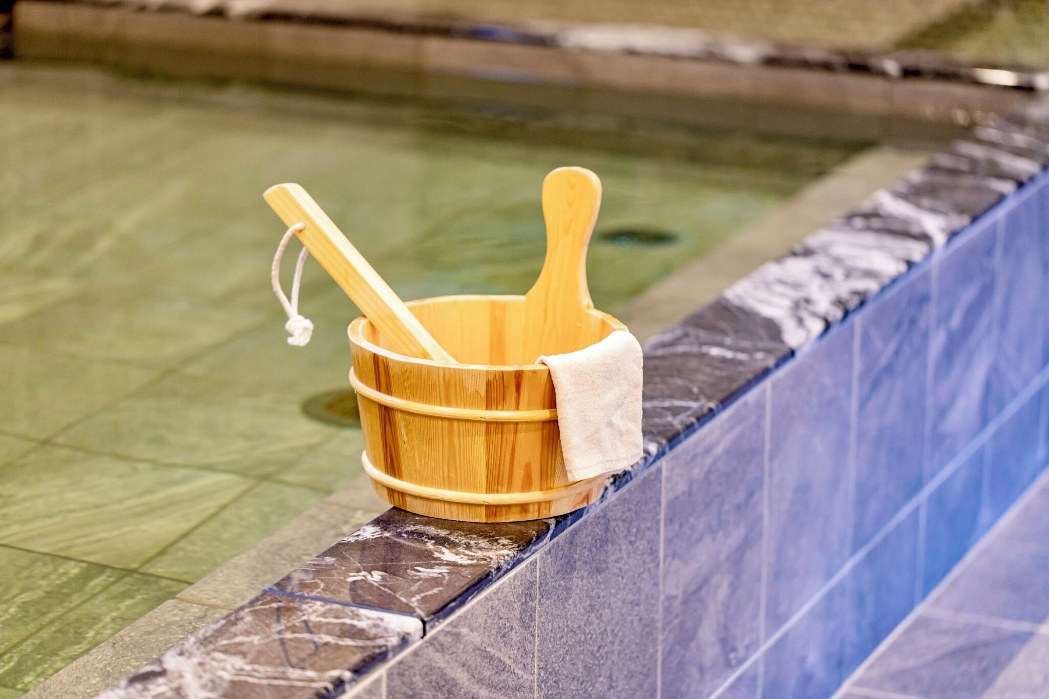 A wooden bucket with a towel and two wooden spoons placed on a tiled pool edge.