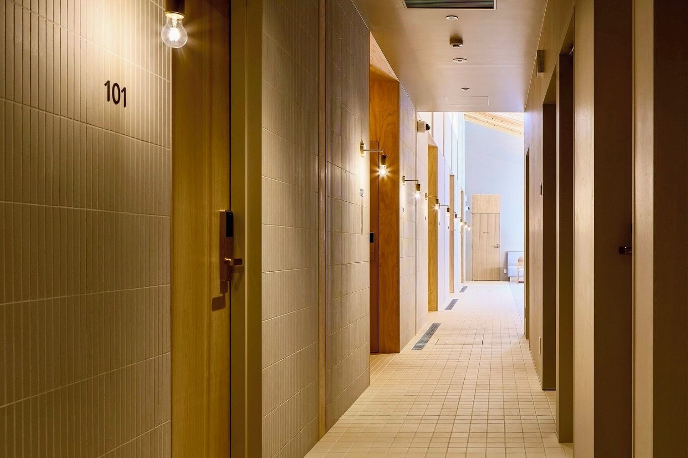 A hotel corridor with numbered doors, a tiled floor, and recessed lighting.