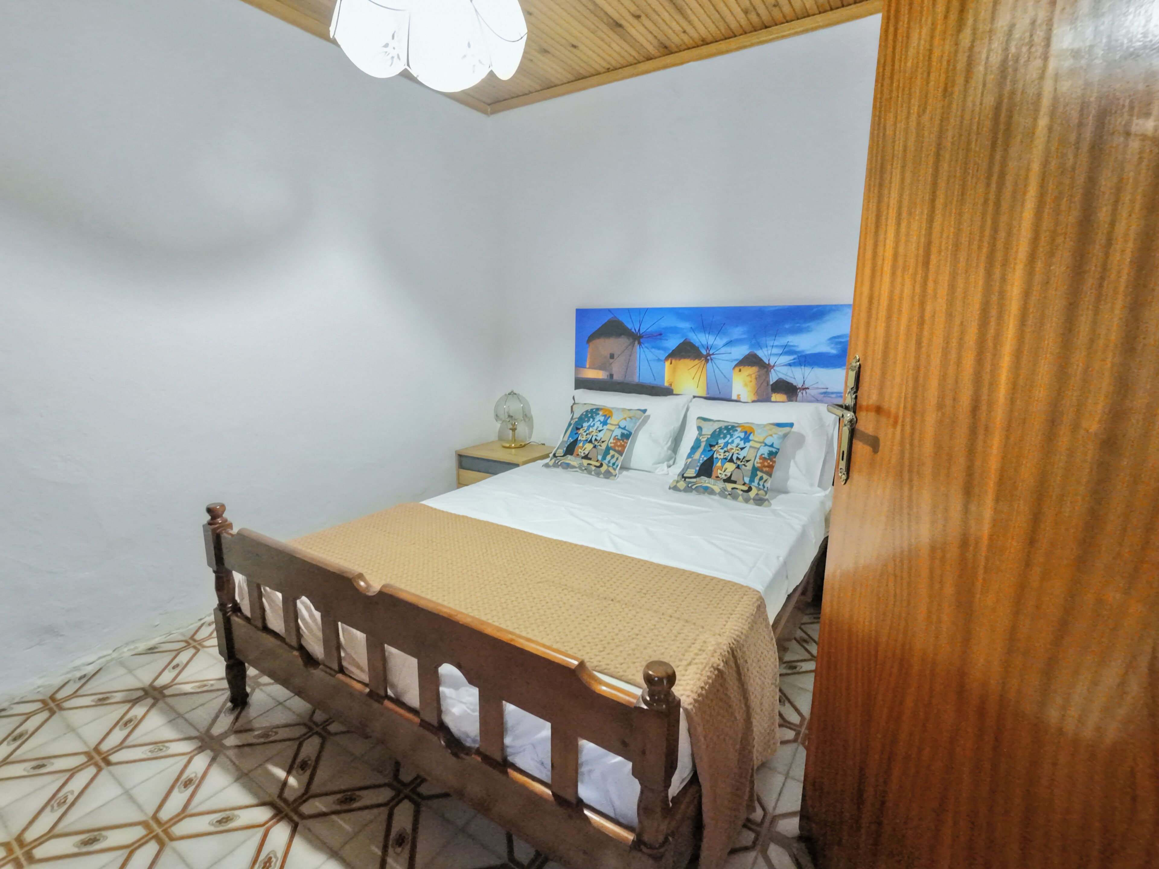 Un dormitorio con cama de madera, mesita de noche, lámpara y un cuadro de molinos en la pared.