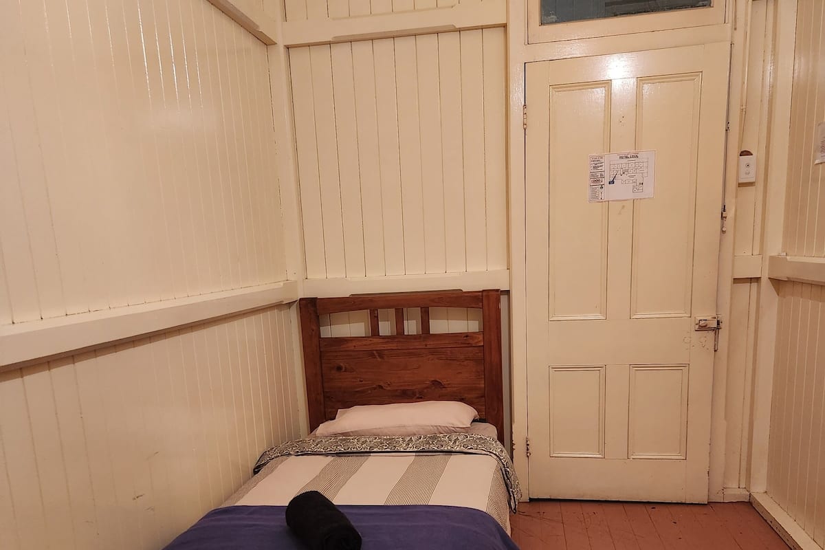 A small, single-bed room with a wooden headboard, a blue towel on the bed, and a door with a window.