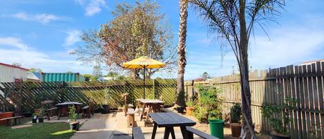 A patio with picnic tables and a yellow umbrella, surrounded by a wooden fence and potted plants.