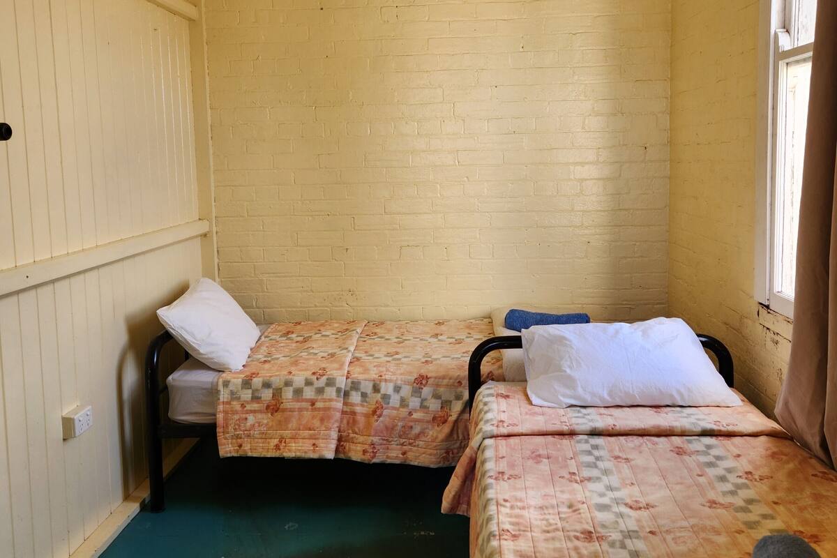 A small, single-room accommodation with two beds, a window, and a wooden wall.