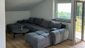 A modern living room with a sectional sofa, a small round table, and a large window with a view. - Charming Alpine Apartment with Sauna (Kobarid)