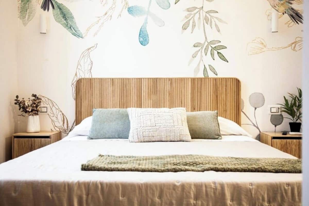 A neatly made bed with a wooden headboard, two pillows, a green throw, and a wall with a nature-themed mural.