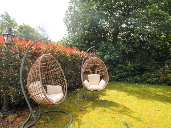 Two wicker egg chairs suspended by metal frames in a garden with green grass and red foliage.