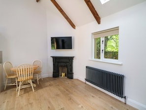 A room with a fireplace, a TV mounted above it, a window with a view of greenery, and a dining area with chairs. - THE MEWS @ DERRYBAWN, pet friendly in Laragh, County Wicklow (Glendalough)