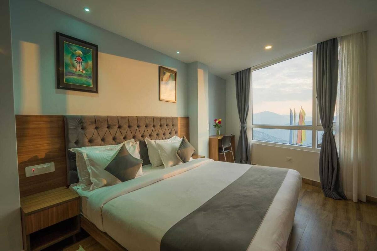 A hotel room with a large bed, a wooden headboard, a desk, and a view of mountains through the window.
