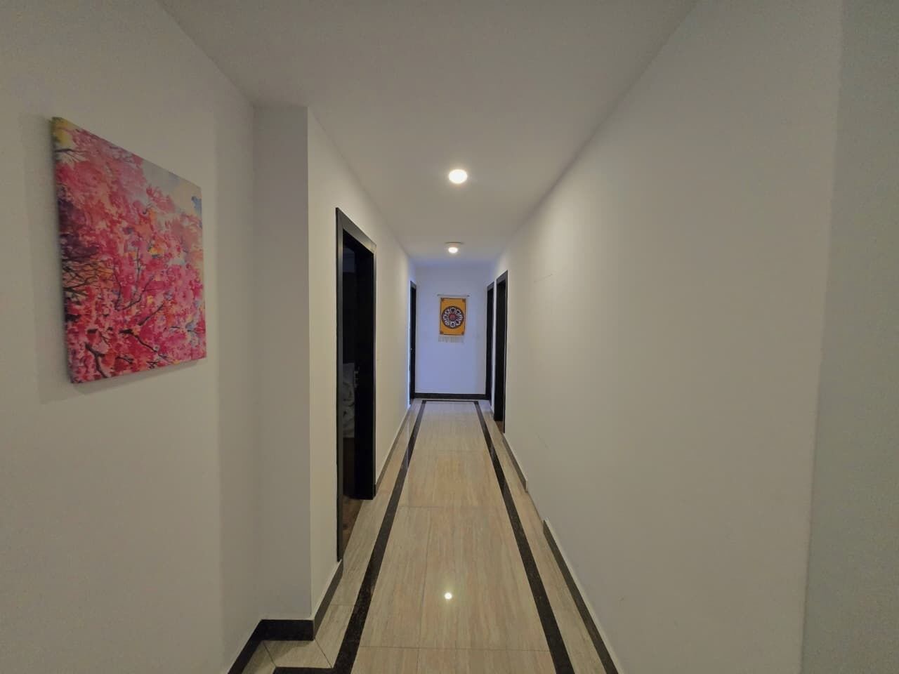 A hallway with a light-colored floor, a painting on the left wall, and a door at the end.