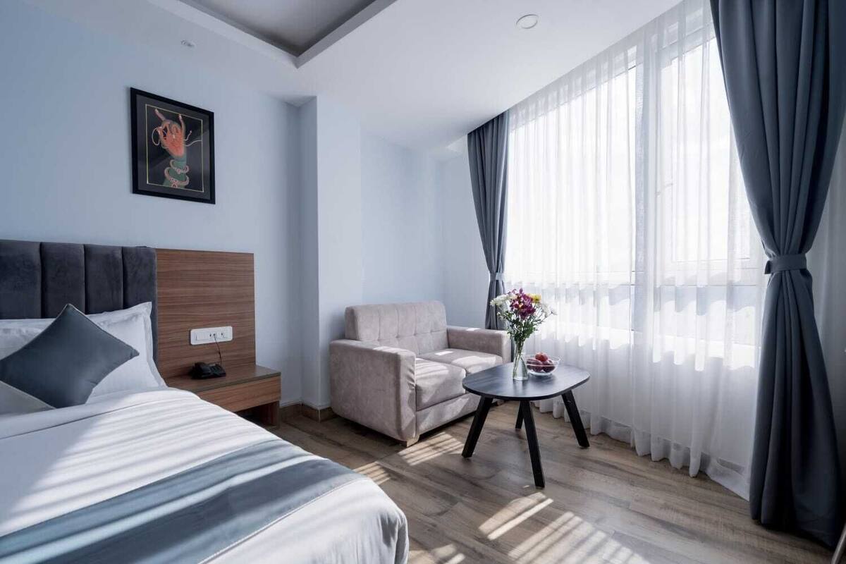 A hotel room with a bed, a chair, a small table with a vase, and a window with curtains.