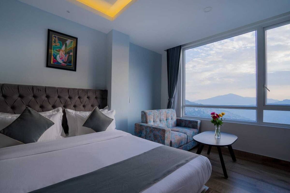 A hotel room with a large bed, a chair, a small table with flowers, a painting on the wall, and a view of the mountains through the window.