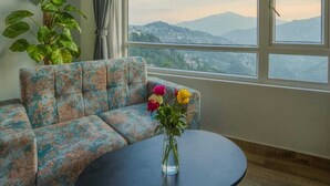 Une pièce avec un canapé, une table ronde, une plante en pot et une vue sur les montagnes, visible à travers de grandes fenêtres.