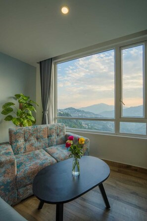 Quarto com sofá, mesa redonda, planta em vaso e vista para montanhas através de amplas janelas.