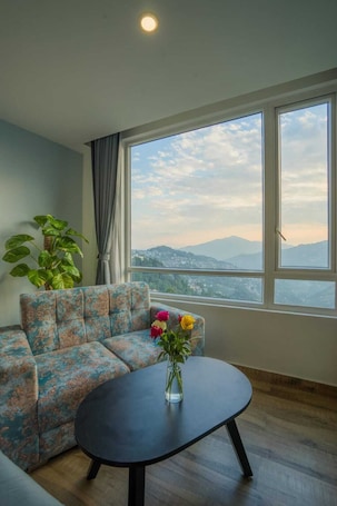 A room with a sofa, a round table, a potted plant, and a view of mountains through large windows.. Mandarava Hotel