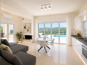 A modern living room with a fireplace, a dining area, and a kitchen with a view of a pool and hills. - Luxury Villa with Private Pool & Stunning Cretan Views Near Heraklion (HERAKLION)