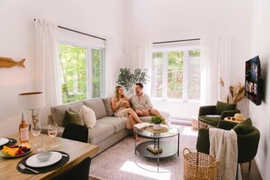 A modern living room with a sofa, a coffee table, a dining table with fruit and wine, and large windows with curtains. - Java with Private Spa, Lake & Kayak (St-Raymond)