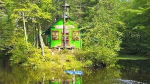 Een groene houten hut met schoorsteen, omringd door bomen en een waterpartij.