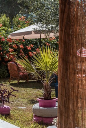 Un jardín con una planta de palmera en una maceta rosa, una silla de mimbre y un paraguas rojo al fondo.