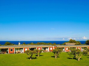 A row of red and white motel buildings with a grassy lawn and trees in front, overlooking a clear blue ocean. - The Apple Garden (Allinge)