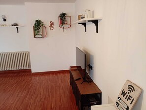 Quarto com piso de madeira, uma TV sobre um suporte, uma escrivaninha e prateleiras com plantas e objetos decorativos.