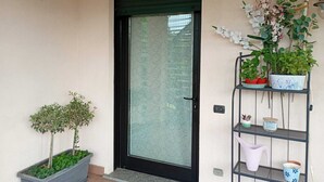 A door with a glass panel, a small potted plant, a metal shelf with various items, and a wall-mounted light fixture. - Bright, spacious apartment between Milan and Como. (Lombardia)