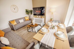 Ein modernes Wohnzimmer mit einer Couch, Couchtisch, Esstisch für vier Personen, einem an der Wand befestigten Fernseher und einer Lampe.