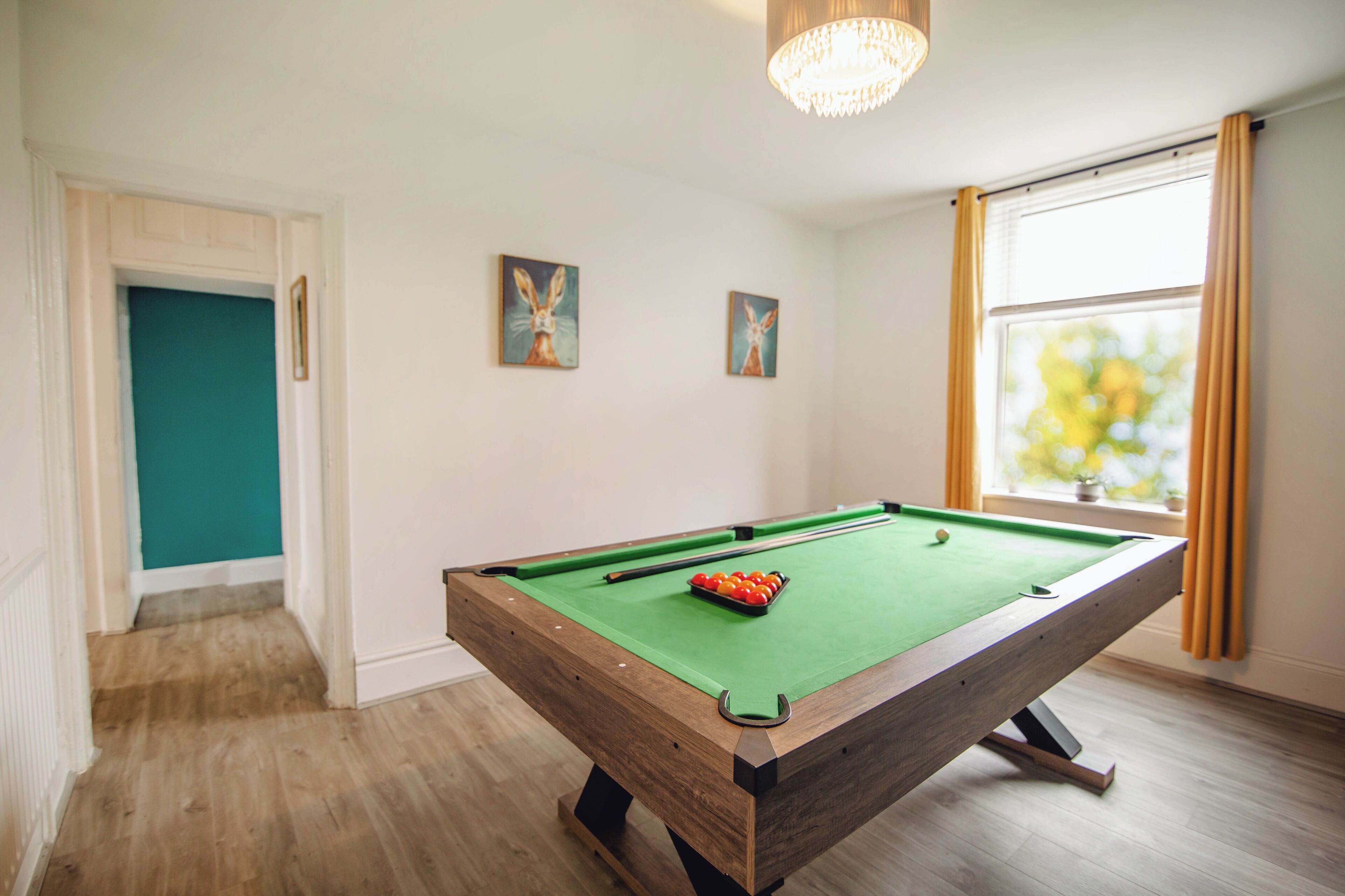 A pool table with balls, a green felt surface, and a cue stick. There are two paintings on the wall and a window with curtains.