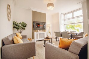 A modern living room with a flat-screen TV, grey sofas, a dining area, and a large window. - Group Haven 5 Bed 2 Bath | Hot Tub | Pool Table (Sheffield)