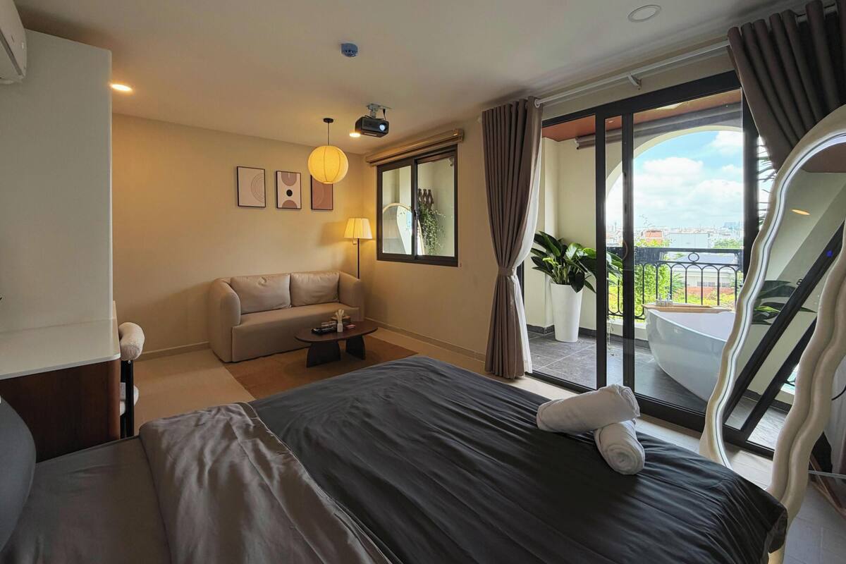 A hotel room with a bed, a sofa, a coffee table, a lamp, a large window, and a balcony with a bathtub.