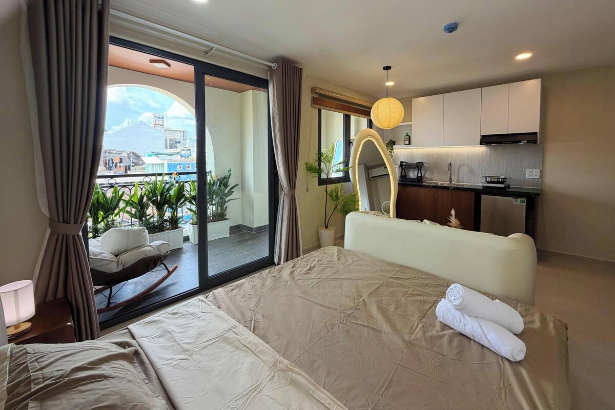 A hotel room with a bed, a view of a balcony with plants, a kitchen area, and a large mirror.
