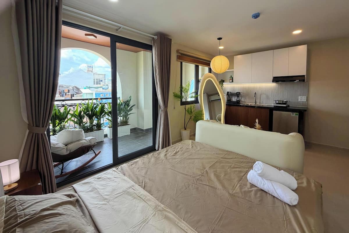 Habitación de hotel con una cama, vista a un balcón con plantas, zona de cocina y un espejo grande.