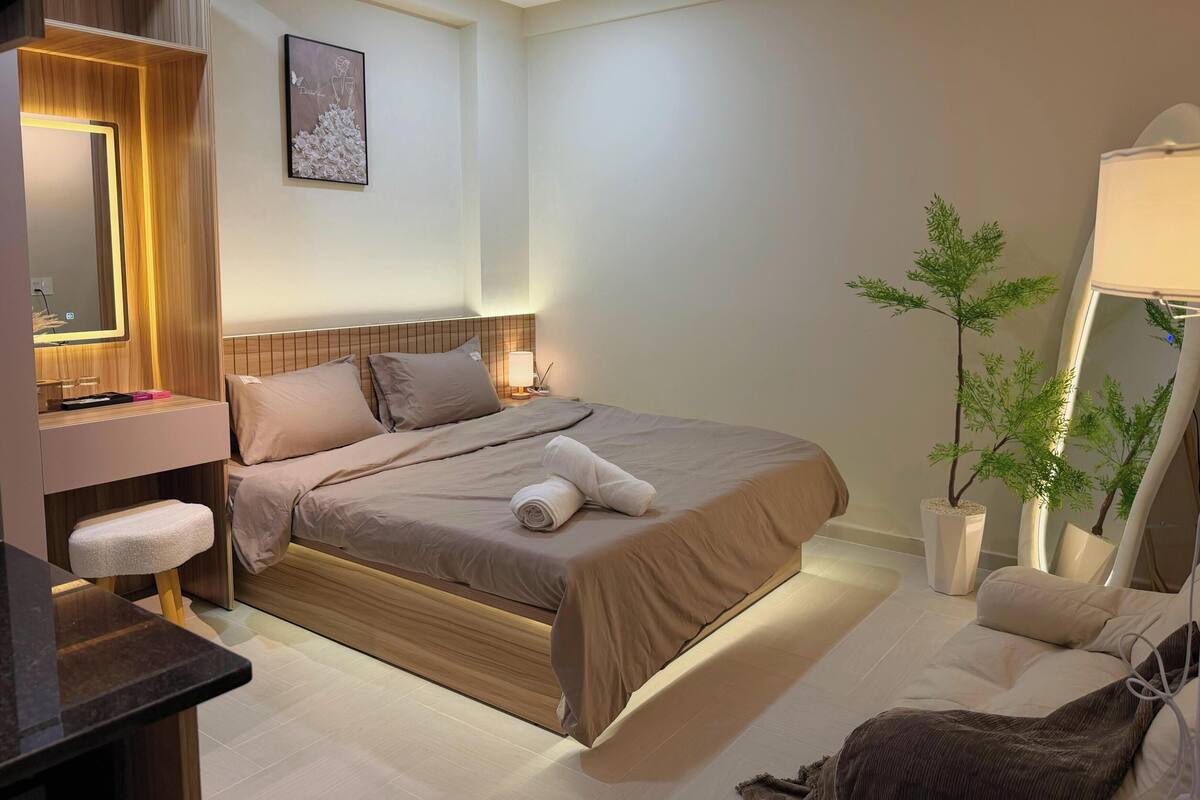 A hotel room with a bed, bedside table, lamp, and a potted plant.