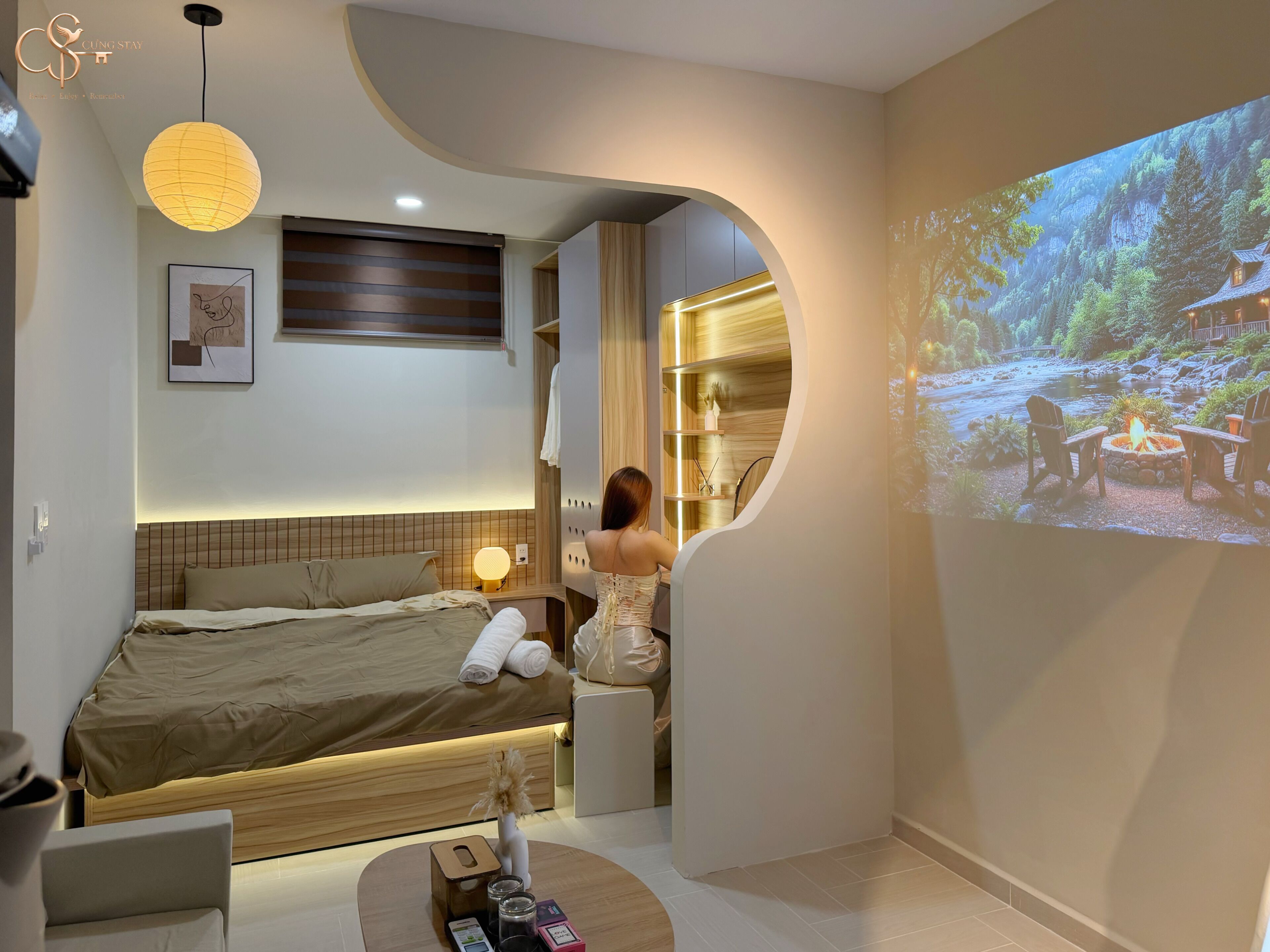 Una habitación de hotel moderna con cama, mesita de noche, una pequeña zona de estar, pantalla de proyección y vistas a un paisaje natural.