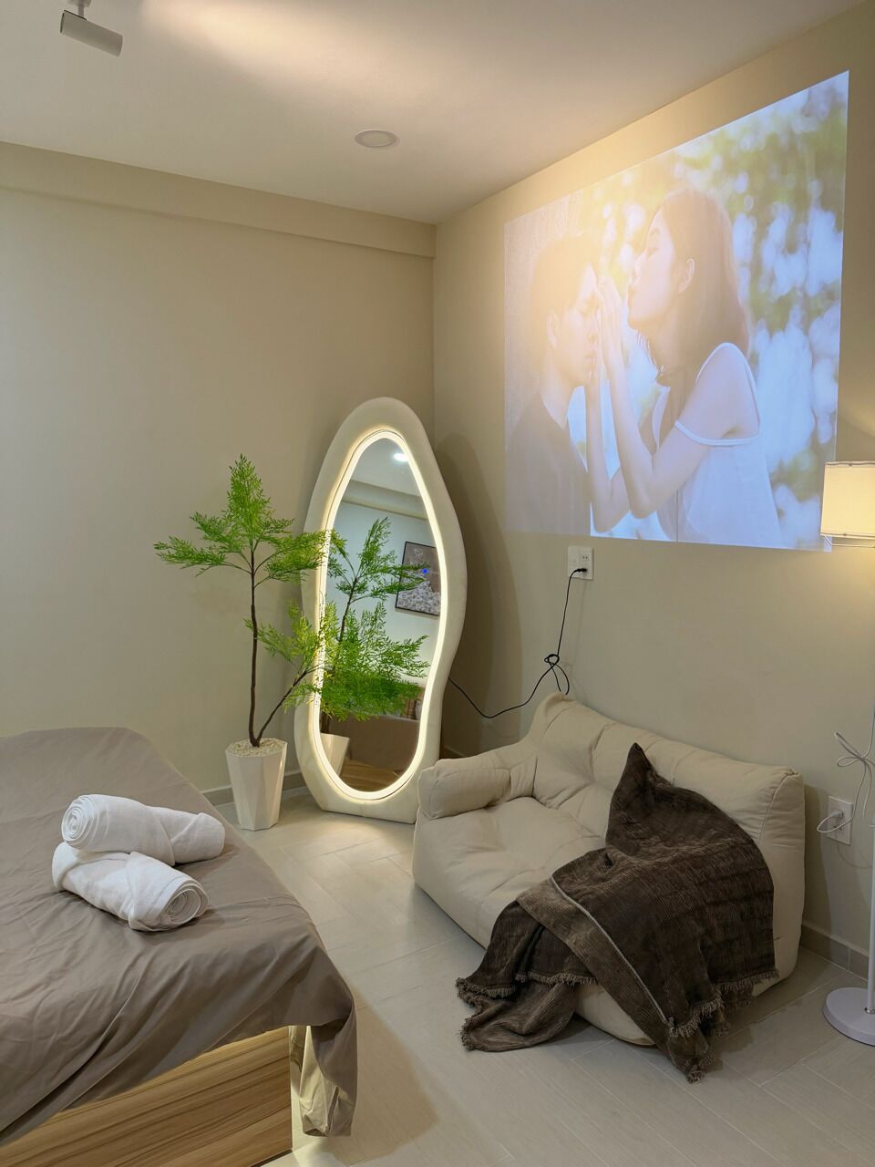 A hotel room with a bed, a sofa, a mirror, a plant, and a wall-mounted TV.