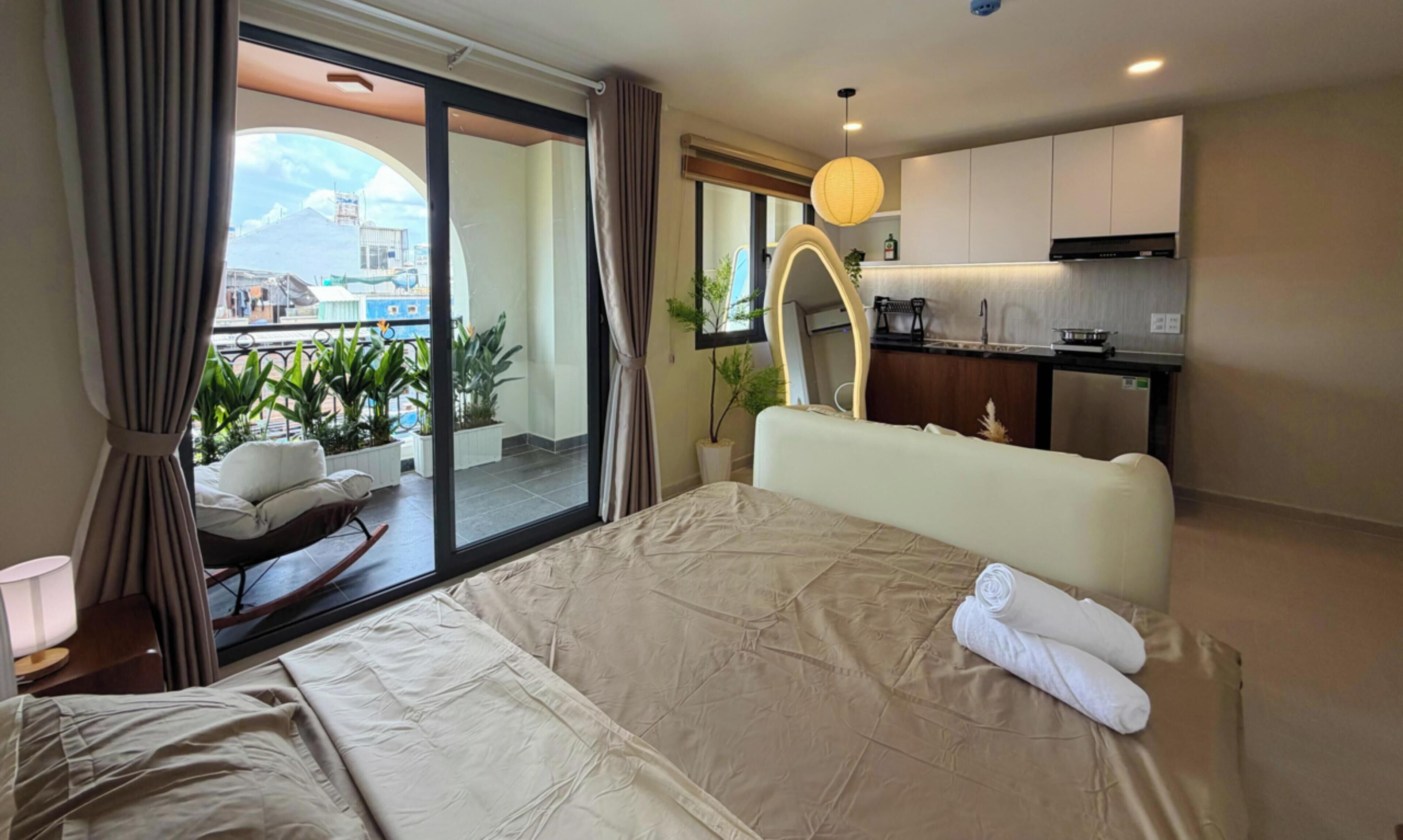 A hotel room with a bed, a small sitting area with a chair and table, a kitchenette, and a balcony with plants.